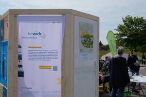 Nahaufnahme der bewirk Ausstellung mit bewirk Logo und Beschreibungstext. Im Hintergrund sind ein Infotisch und Menschen zu sehen